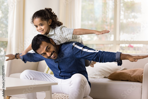 Obraz na plátne Joyful excited little Indian daughter girl riding dads back with flying open arms, playing airplane with strong father at home, laughing, giggling, smiling