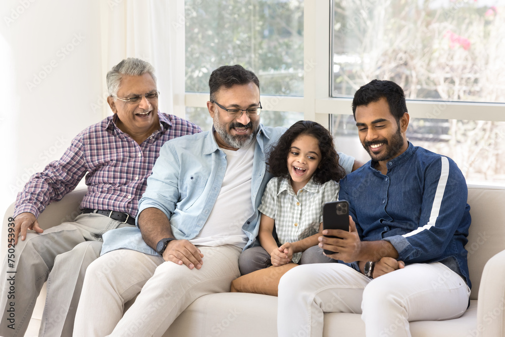 Cheerful Indian great grandpa, grandad, father and kid using mobile phone together, resting on home couch, talking on video call, enjoying family Internet communication Cheerful Indian great grandpa, grandad, father and kid using mobile phone together, resting on home couch, talking on video call, enjoying family Internet communication