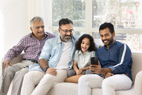 Cheerful Indian great grandpa, grandad, father and kid using mobile phone together, resting on home couch, talking on video call, enjoying family Internet communication