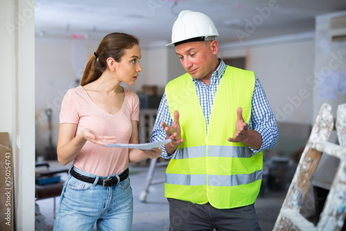 Frustrated young adult female designer arguing with male worker while examining indoor construction site at renovating object