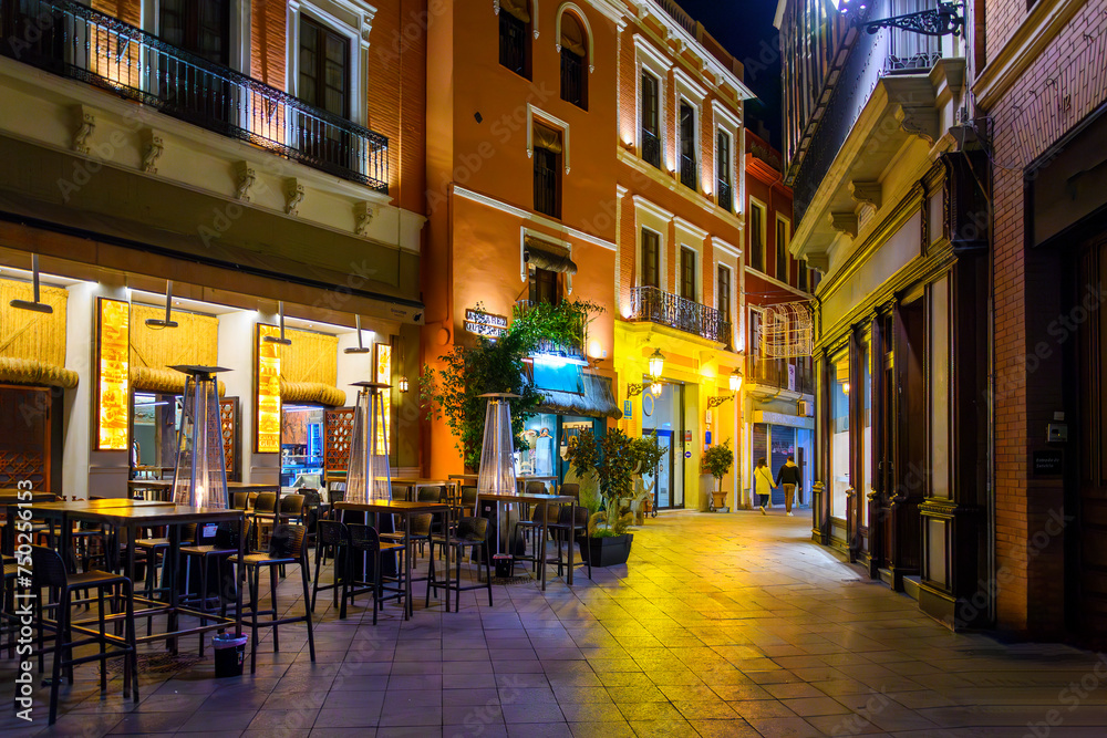 Cafes and shops colorfully illuminated at night in the central Barrio Santa Cruz district of the Andalusian city of Seville, Spain.