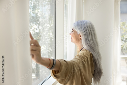 Happy inspired senior Latin woman opening white transparent veils, enjoying morning daylight, looking out of large window, smiling, relaxing in comfortable apartment. Side view,