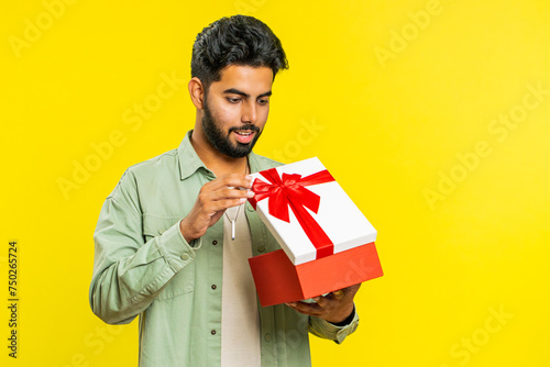 Happy Indian young man opening birthday gift box with red ribbon. Holidays surprise concept. Smiling Arabian bearded guy receive great wrapped present celebrating isolated on yellow studio background