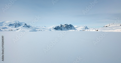 Winter Antarctica snow covered nature landscape. Frozen ocean, mountain range in background, sunny weather, blue sky. Desert white land of ice. Polar arctic travel scene. Low angle drone flight photo