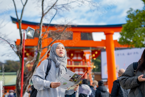 Travel, muslim travel, Asian Muslim female tourists visitor learning about history of fushimi inari shrine in travel book while walking through senbon torii path in Kyoto Japan.