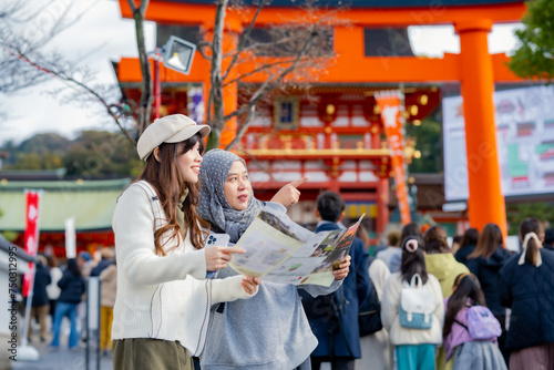 Travel, muslim, Two Asian female tourists of different religions friends visitor learning about history of fushimi inari shrine in travel book while walking through senbon torii path in Kyoto Japan. photo
