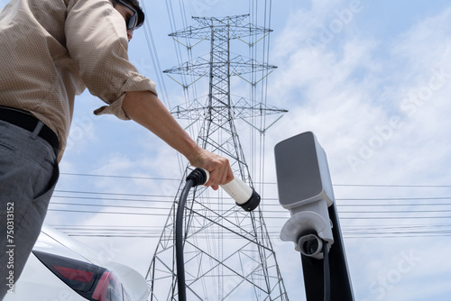 Man recharge EV electric car battery at charging station connected to electrical power grid tower on sky background as electrical industry for eco friendly vehicle utilization. Expedient photo