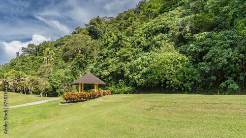 The green lawn is neatly trimmed. A footpath winds between the hills. A decorative gazebo with a conical roof stands by the side of the road. Lush tropical vegetation against a blue sky and clouds.