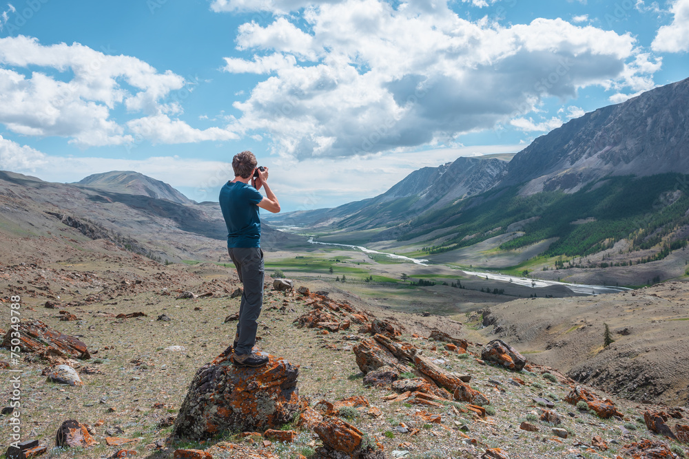 Man shoots landscape with mountain river in alpine valley. Guy with photo camera on stone with view to long river in big mountains. Conifer trees in wide valley among rocky stony hills and steep crags