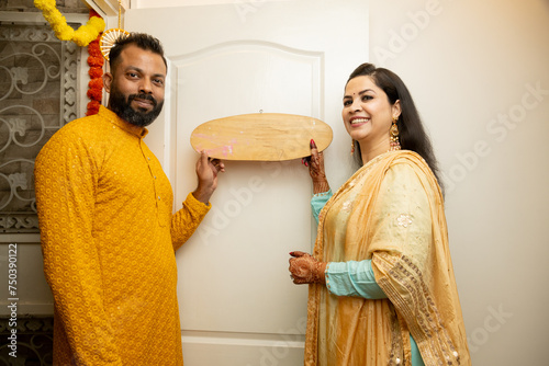 Happy indian couple of standing in their new house with name plate in hand. concept of new festival purchase.