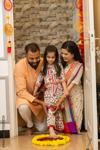 Happy indian family of three doing grah pravesh ritual or entering new house for the first time. daughter entering house with right feet stepping in a plate filled liquid kumkum. Griha Pravesh Concept photo