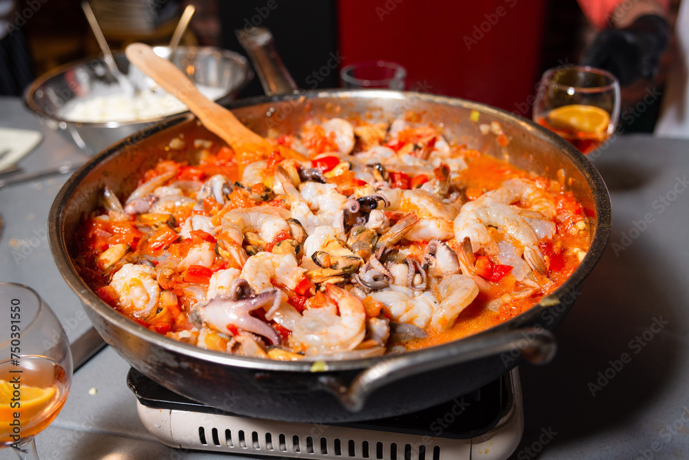 Seafood Paella Cooking in a Large Pan. A large pan filled with seafood paella featuring shrimp and squid, simmering on a stove, with the vibrant colors of fresh ingredients on display.