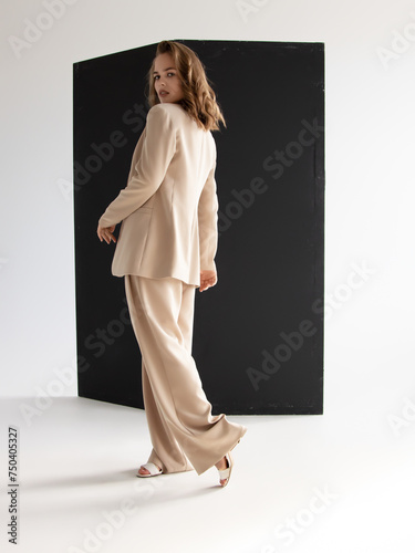 Young brunette curly woman in beige suit. Elegant style. A beautiful fashion model poses in a loose beige pantsuit. Female fashion. Full-length studio portrait.