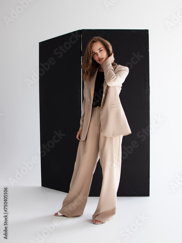 Young brunette curly woman in beige suit. Elegant style. A beautiful fashion model poses in a loose beige pantsuit. Female fashion. Full-length studio portrait.