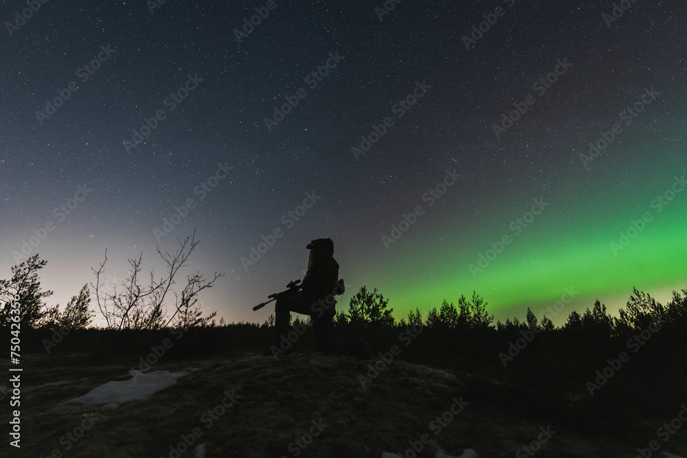 A military soldier with a night vision device and a rifle with a silencer is sitting in the forest against the background of the starry sky and northern lights.