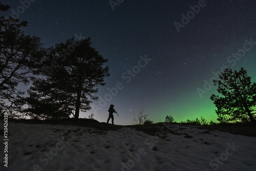 A soldier with a night vision device and a rifle with a silencer in the forest against the background of the starry sky and northern lights.