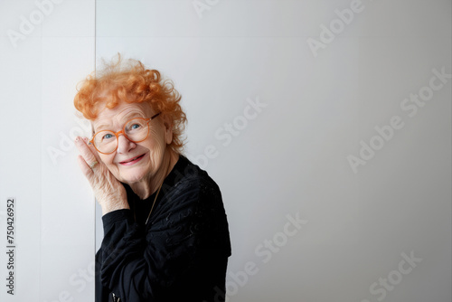 A cheerful happy ederly woman l with gray hair looks out from behind a white wall. Hide and seek games. Happy mood. A cute old woman. The woman looks at the camera and smiles. banner. Place for text.
