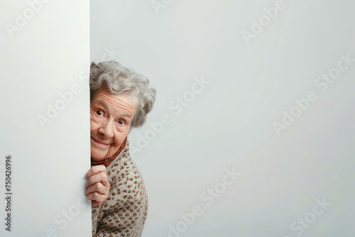 A cheerful happy ederly woman l with gray hair looks out from behind a white wall. Hide and seek games. Happy mood. A cute old woman. The woman looks at the camera and smiles. banner. Place for text. photo