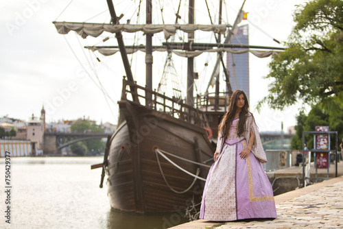 Obraz na plátně Young woman, Hispanic, beautiful, brunette, with an elegant violet vintage dress, posing by the river with a replica of Spanish galleon in the background