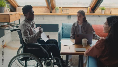 Medium shot of African American man with disability sitting in wheelchair in loft coworking office at project team meeting, proposing new business idea and showing graphs, female colleagues listening photo