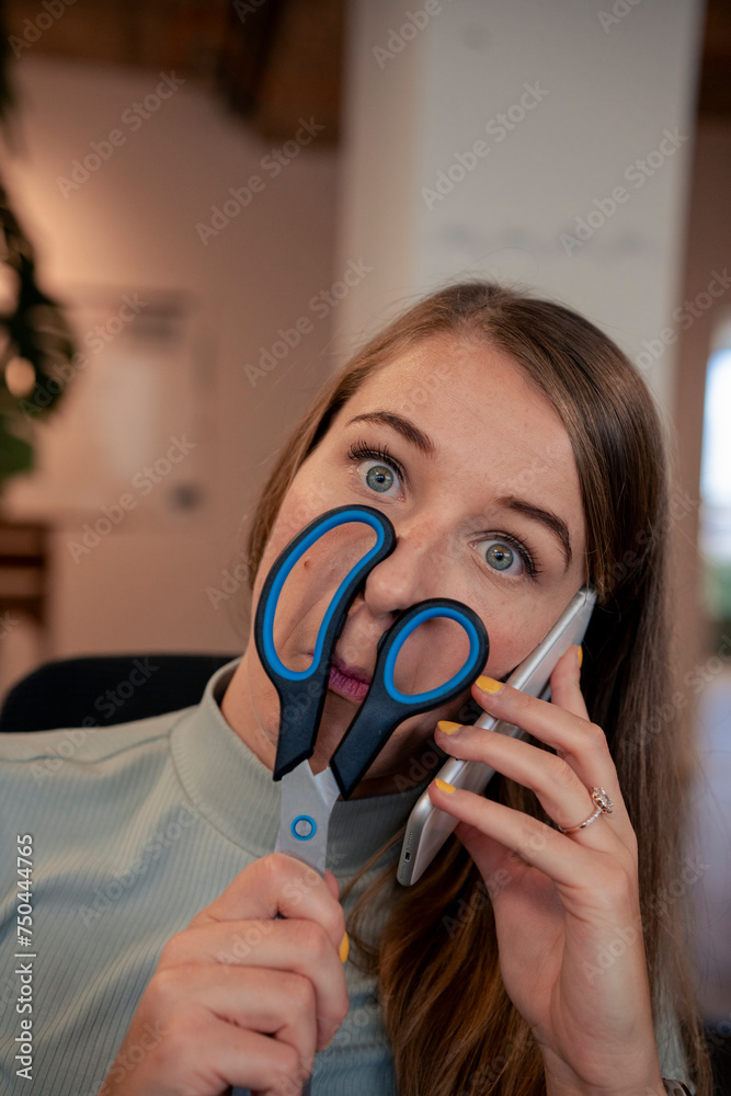 Playful young businesswoman holding scissors over face and talking on smart phone in office Playful young businesswoman holding scissors over face and talking on smart phone in office