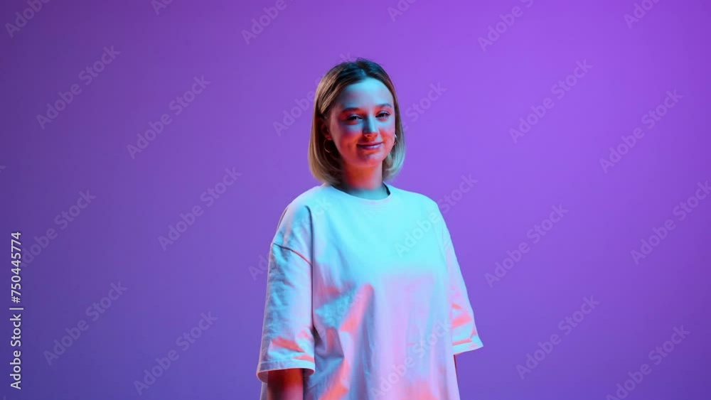 Beautiful young girl in white t-shirt standing, posing with smile against purple studio background in neon light. Video, 4k. Concept of youth, human emotions, casual fashion, lifestyle Beautiful young girl in white t-shirt standing, posing with smile against purple studio background in neon light. Video, 4k. Concept of youth, human emotions, casual fashion, lifestyle