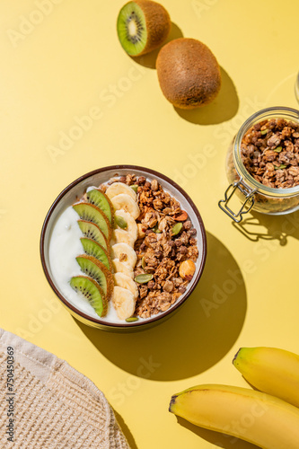 Homemade granola with banana, kiwi and nuts in a bowl on a yellow background with fresh fruits.