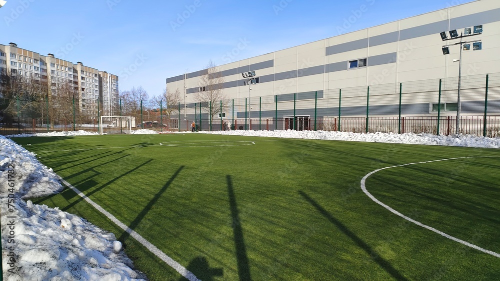 The stadium is located among various buildings and trees. The soccer field with goals and artificial grass is cleared of snow. Pillars with lighting lanterns are installed around it. Sunny weather The stadium is located among various buildings and trees. The soccer field with goals and artificial grass is cleared of snow. Pillars with lighting lanterns are installed around it. Sunny weather