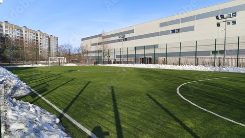 The stadium is located among various buildings and trees. The soccer field with goals and artificial grass is cleared of snow. Pillars with lighting lanterns are installed around it. Sunny weather