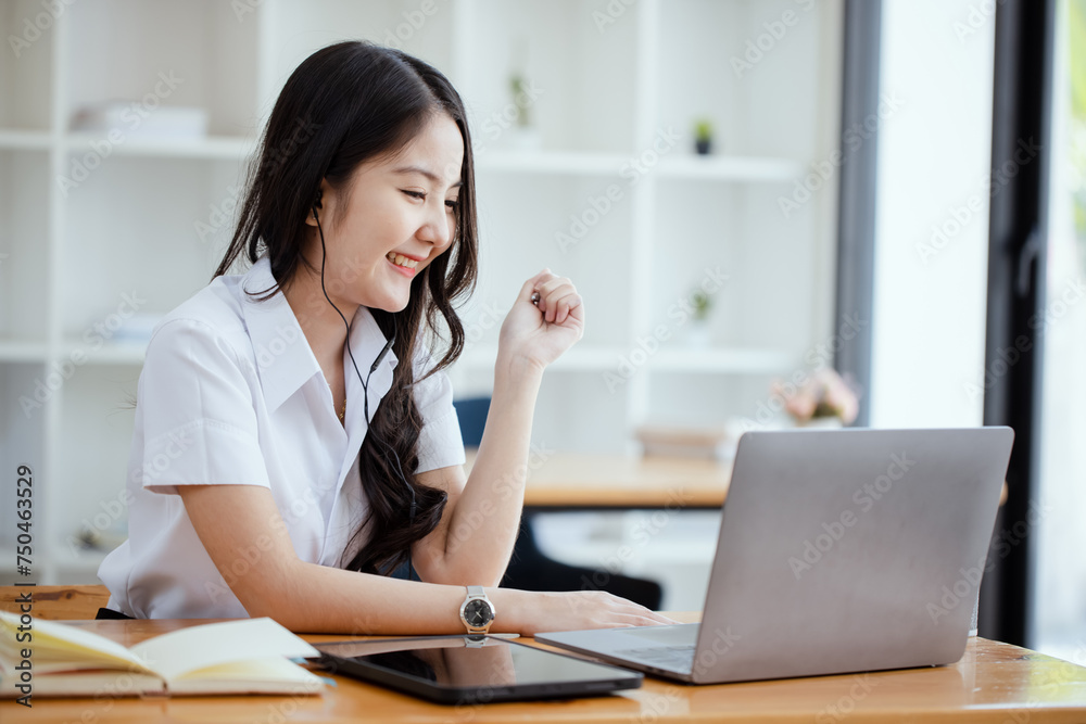 Happy young asian woman wearing earphones and using laptop computer at desk in office, Female asian student with laptop computer, College female student learning online. Happy young asian woman wearing earphones and using laptop computer at desk in office, Female asian student with laptop computer, College female student learning online.