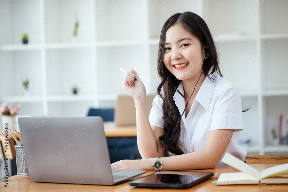 Happy young asian woman wearing earphones and using laptop computer at desk in office, Female asian student with laptop computer, College female student learning online. Happy young asian woman wearing earphones and using laptop computer at desk in office, Female asian student with laptop computer, College female student learning online.
