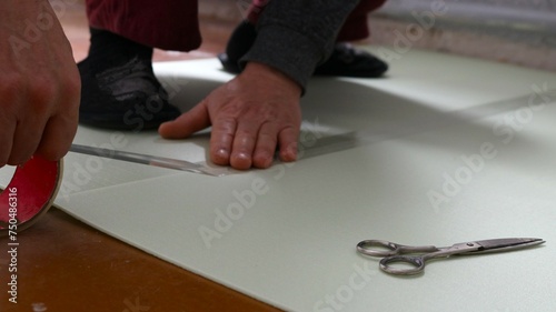 male hands glue together with transparent tape sheets of soft light-colored underlay for laminate on the floor and scissors lying nearby, do it yourself preparing the flooring at home
