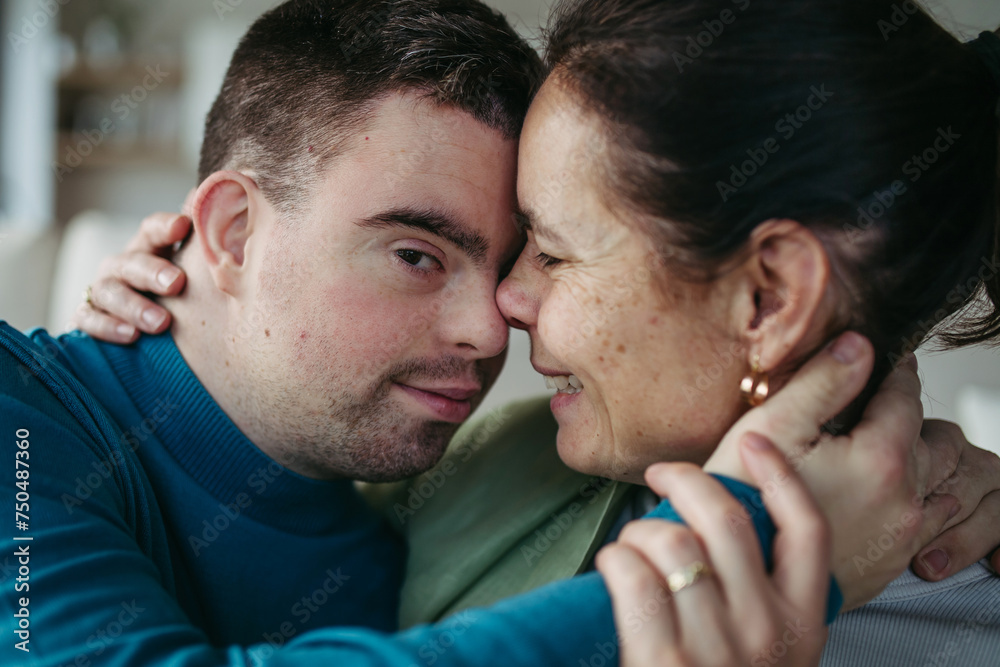 Fototapeta premium Portrait of young man with Down syndrome with his mother at home, holding, touching with foreheads. Concept of love and parenting disabled child. Banner with copyspace.