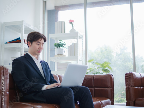 Business man portrait Asian one person wearing blue suit sitting on sofa looking serious and determined expression hand holding laptop notebook ready for working online sale inside the home office