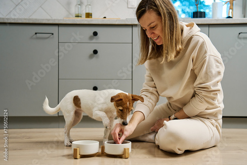 Woman putting food bowl with feed for her dog on the floor in kitchen, Female owner spending time together with pet at home, Animal feeding and pet care photo