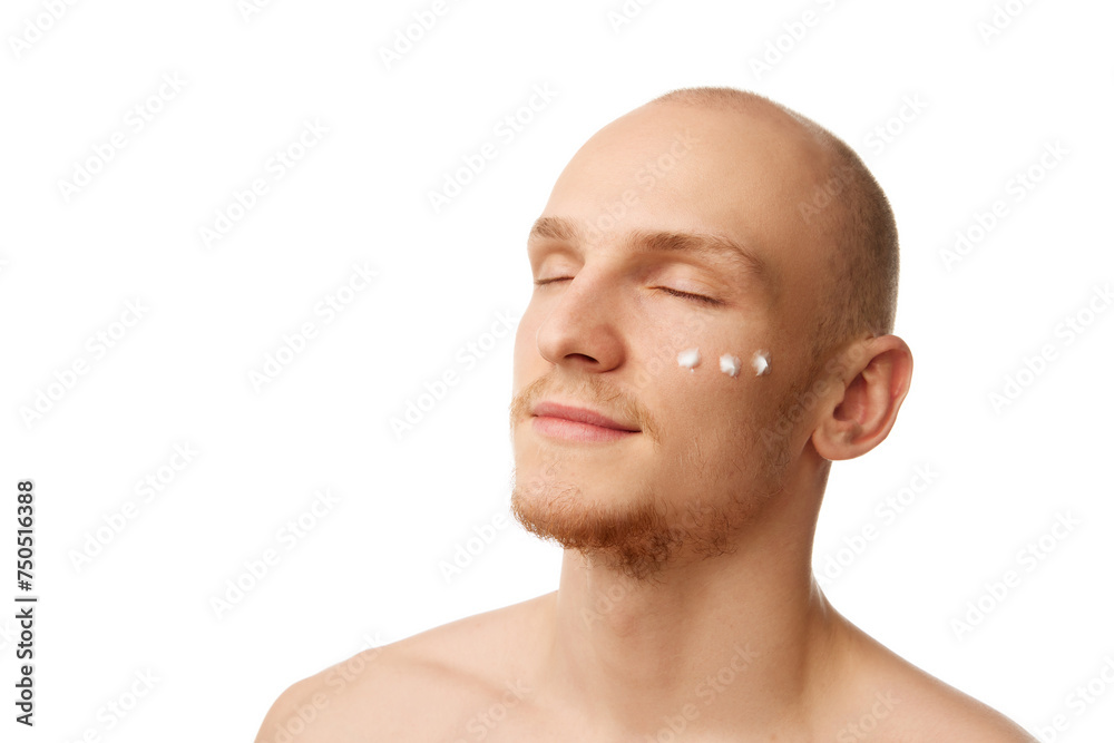 Fototapeta premium Portrait of handsome bald young man standing with eyes closed with moisturizing cream on face isolated over white studio background. Concept of skin care, male beauty and cosmetics, youth, self-care