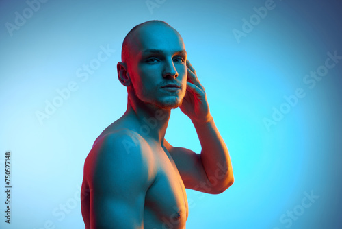 Portrait of handsome young bald man posing shirtless, looking at camera against blue studio background in neon light. Concept of male beauty, sportive and healthy lifestyle, body care