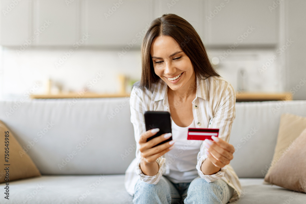 Happy young european woman sitting on couch, using smartphone and credit card Happy young european woman sitting on couch, using smartphone and credit card