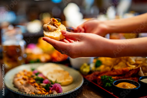 A close-up of hands holding a small open-faced sandwich topped with assorted toppings. In the background, an out-of-focus table set with various dishes, including more sandwiches and dipping sauces.