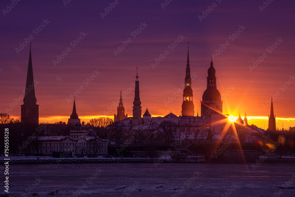 Naklejka premium A beautiful, colorful sunrise landscape with city centre building silhouette against bright morning sky. A winter lansdcape with frozen river Daugava in Latvia capital Riga.