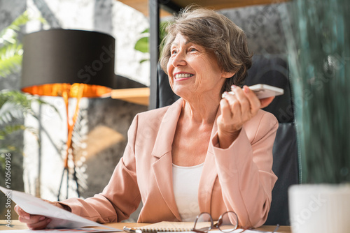 Slika na platnu Happy caucasian senior manager ceo businesswoman using cellphone to record voice message, dealing with papers documents at office