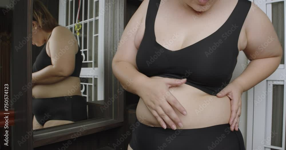 Asian oversize woman in front of a mirror looks at her body. Excess belly fat, Unhealthy lifestyle, Diet concept.