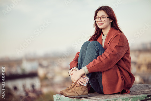 Happy and satisfied teenage girl in glasses with long red hair in red coat sits with his knees bent and looks away, against the background of sky and blurred cityscape. © TanyaJoy