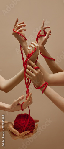 Unity. Woman's hand holds ball of red threads and other hands intertwined with each other tied with this thread against sandy color studio background. Concept of human touch, beauty and care.