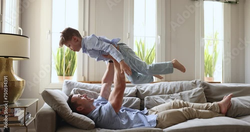 Cheery father plays with little son, lifting lovely kid on outstretched hands, enjoy playtime in cozy living room. Preschooler boy have fun with dad, imagining flying in the air, pretend to be a plane photo