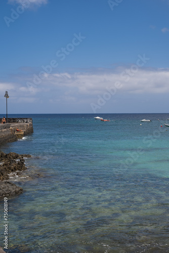 Seascape. Dock of the village of Arrieta with group of boats anchored near the dock in the background. Turquoise Atlantic Ocean. Big white clouds. Village of Arrieta. Lanzarote, Canary Islands, Spain © Jess