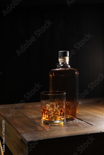 Whiskey with ice cubes in glass and bottle on wooden crate against black background © New Africa