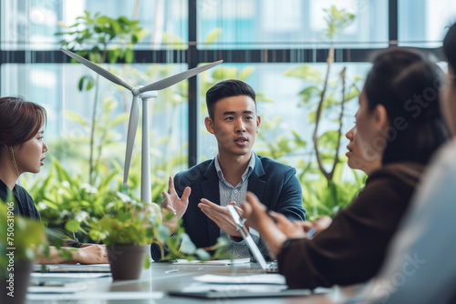 professional of East Asian ethnicity gesture towards a small model of a wind turbine and a solar cell array placed on the meeting table