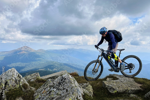 Cyclist man riding electric mountain bike outdoors. Male tourist biking along grassy trail in the mountains, wearing helmet and backpack. Concept of sport, active leisure and nature.