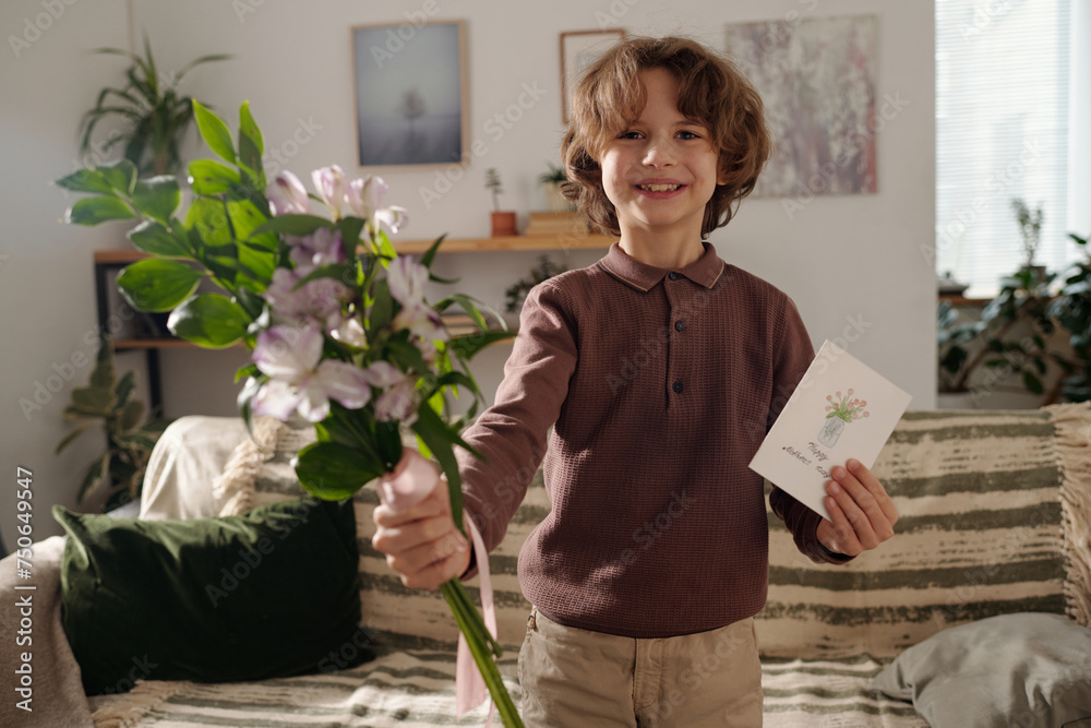 Cute cheerful boy with handmade postcard for mother day passing bunch of flowers and looking at camera while standing against couch Cute cheerful boy with handmade postcard for mother day passing bunch of flowers and looking at camera while standing against couch